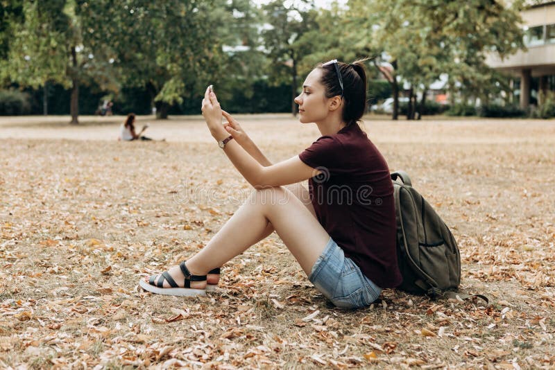 Een meisje met een rugzak zit in een de herfstpark en gebruikt een celtelefoon royalty-vrije stock foto Een meisje met een rugzak zit in een de herfstpark en gebruikt een celtelefoon royalty-vrije stock foto