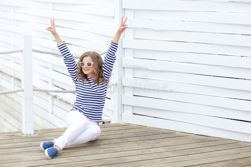 Een meisje in witte strakke jeans en sportenschoenen die tegen een witte houten muur stellen In een zeemanst-shirt en glazen krul royalty-vrije stock foto's Een meisje in witte strakke jeans en sportenschoenen die tegen een witte houten muur stellen In een zeemanst-shirt en glazen krul royalty-vrije stock foto's
