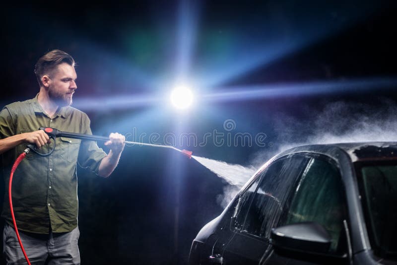 Een mens met een baard of autowasmachine wast een grijze auto met een hoge drukapparaat bij nacht in een autowasserette stock afbeeldingen Een mens met een baard of autowasmachine wast een grijze auto met een hoge drukapparaat bij nacht in een autowasserette stock afbeeldingen