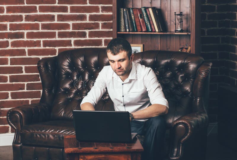 Een mens zit op een luxueuze bank en werkt achter laptop in zijn bureau aan de achtergrond van het boekenrek stock fotografie Een mens zit op een luxueuze bank en werkt achter laptop in zijn bureau aan de achtergrond van het boekenrek stock fotografie