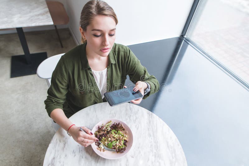 Een mooi meisje maakt een foto van een salade in een restaurant Blogger neemt voedsel op de lijst dichtbij het venster op royalty-vrije stock fotografie Een mooi meisje maakt een foto van een salade in een restaurant Blogger neemt voedsel op de lijst dichtbij het venster op royalty-vrije stock fotografie
