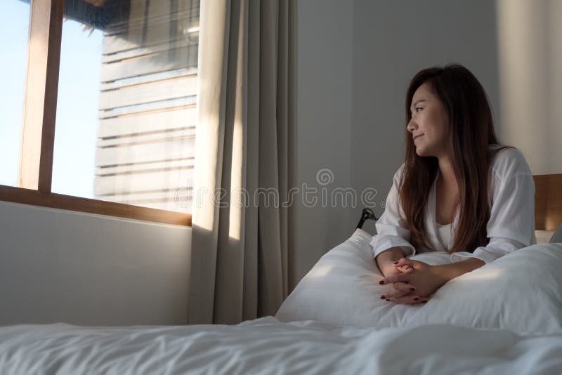 Een mooie Aziatische vrouwenzitting op het bed en het kijken buiten het venster stock fotografie Een mooie Aziatische vrouwenzitting op het bed en het kijken buiten het venster stock fotografie