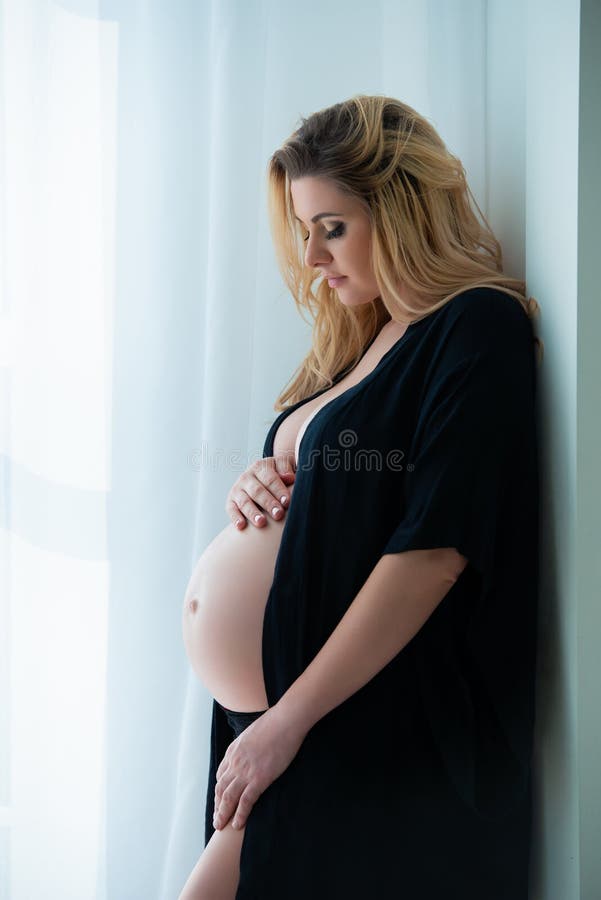 A beautiful pregnant woman in a black robe stands near a bright window. Feminine sexuality, femininity and beautiful pregnancy. A beautiful pregnant woman in a black robe stands near a bright window. Feminine sexuality, femininity and beautiful pregnancy.