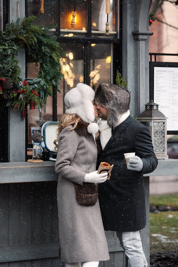 Fashion couple of lovers standing on winter street in snowy weather, men and women wear fur hat and warm wool coat, hugging, drinking hot drinks. St. Valentines Day, Christmas holidays concept. Fashion couple of lovers standing on winter street in snowy weather, men and women wear fur hat and warm wool coat, hugging, drinking hot drinks. St. Valentines Day, Christmas holidays concept.
