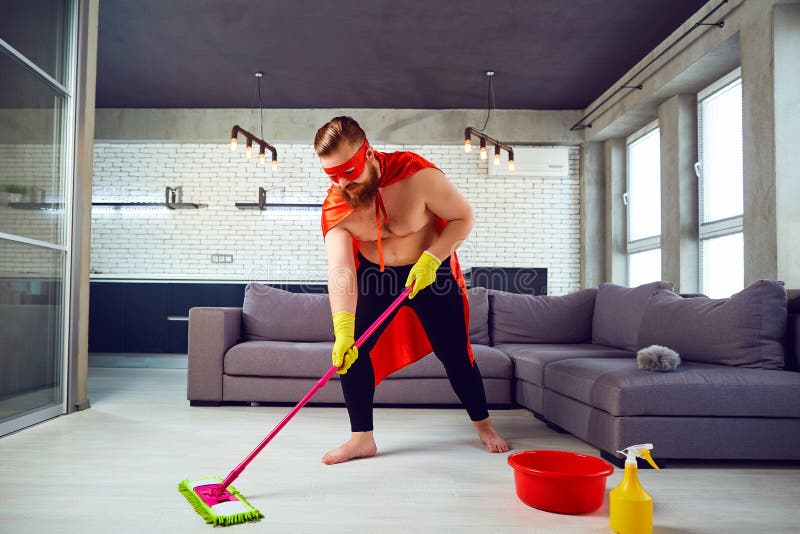 Een vette, grappige mens in een superherokostuum maakt het huis schoon stock afbeelding Een vette, grappige mens in een superherokostuum maakt het huis schoon stock afbeelding