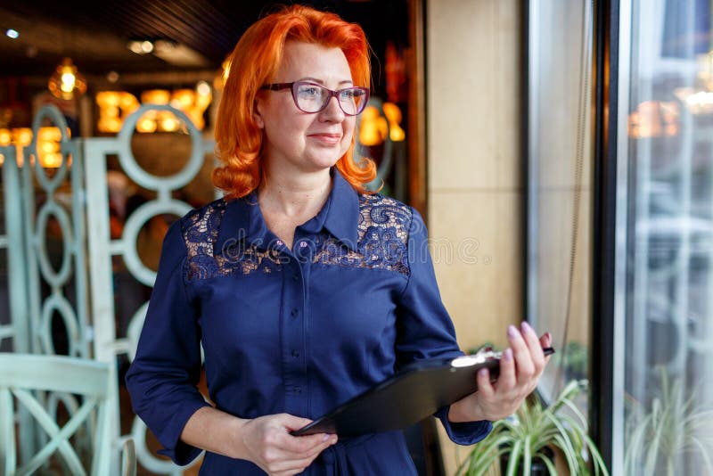 Een vrouw met rood haar, tribunes dichtbij het venster en onderzoekt de afstand, die een klembord houden stock afbeelding Een vrouw met rood haar, tribunes dichtbij het venster en onderzoekt de afstand, die een klembord houden stock afbeelding