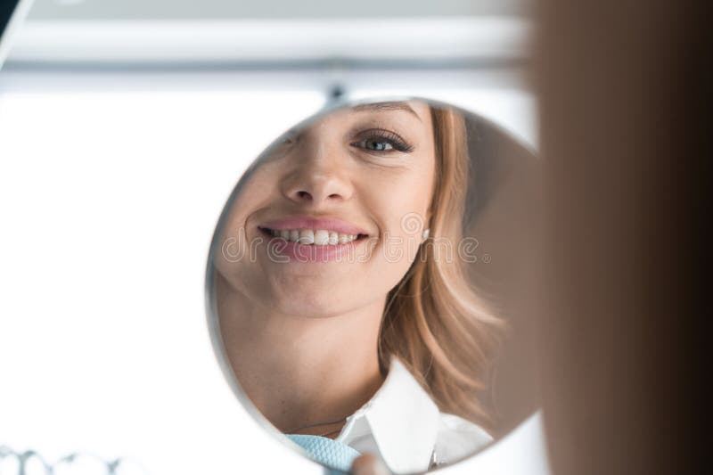 Woman in dental chair happily holding mirror, looking at the wonderful result of the dentist&#x27;s work and enjoying her charming smile. High quality photo. Woman in dental chair happily holding mirror, looking at the wonderful result of the dentist&#x27;s work and enjoying her charming smile. High quality photo