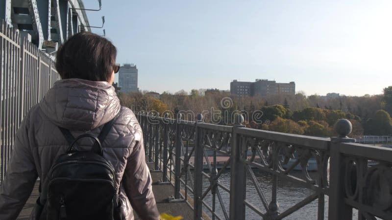 Een vrouw van middelbare leeftijd loopt alleen met een boeket met gele bladeren op een brug over een rivier op een zonnige dag ..