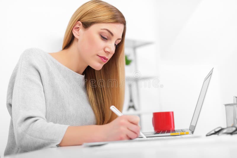 Een vrouw in een wit bureau zit bij een lijst en het schrijven Een jonge dame die laptop met behulp van vult documenten in stock afbeeldingen Een vrouw in een wit bureau zit bij een lijst en het schrijven Een jonge dame die laptop met behulp van vult documenten in stock afbeeldingen
