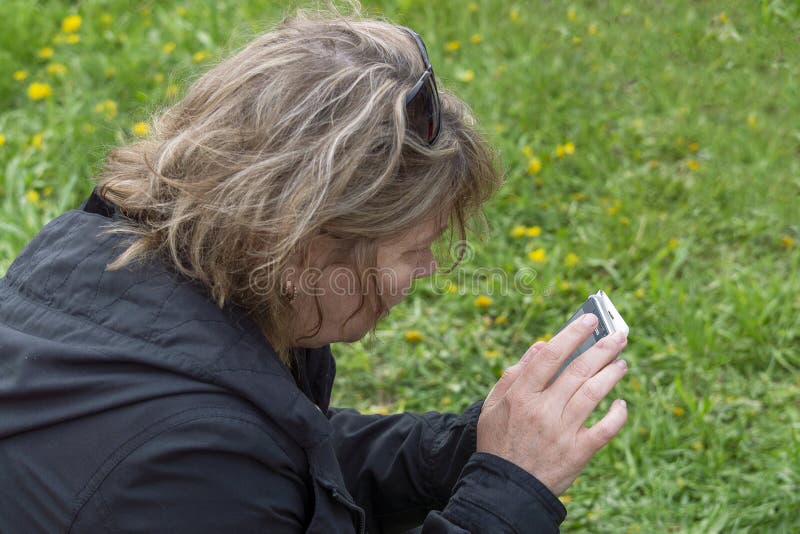 Een vrouw zit op een groen gras en onderzoekt het smartphone, achter en zijaanzicht royalty-vrije stock foto Een vrouw zit op een groen gras en onderzoekt het smartphone, achter en zijaanzicht royalty-vrije stock foto