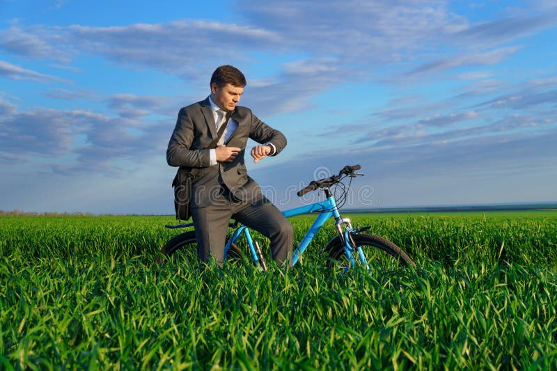 Een zakenman rijdt een fiets met een aktetas en controleert zijn polshorloge op een groen grijs veld dat in een pak is gekleed