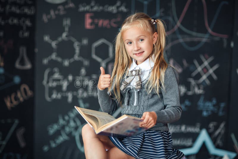 Een zoet meisjesschoolmeisje zit op een bord met de achtergrond van schoolformules Het de werkenthuiswerk door een boek te lezen royalty-vrije stock foto's Een zoet meisjesschoolmeisje zit op een bord met de achtergrond van schoolformules Het de werkenthuiswerk door een boek te lezen royalty-vrije stock foto's