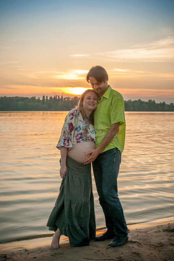 Een zwangere mooie vrouw met haar echtgenoot op het strand die van de rivierbank en wat betreft haar buik met liefde en zorg glim royalty-vrije stock foto Een zwangere mooie vrouw met haar echtgenoot op het strand die van de rivierbank en wat betreft haar buik met liefde en zorg glim royalty-vrije stock foto