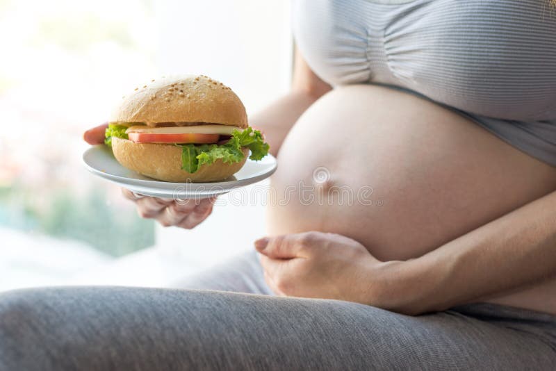 Een zwangere vrouw met buik door het venster die een hamburger houden Concept snel voedsel en het ongezonde eten tijdens zwangers royalty-vrije stock fotografie Een zwangere vrouw met buik door het venster die een hamburger houden Concept snel voedsel en het ongezonde eten tijdens zwangers royalty-vrije stock fotografie