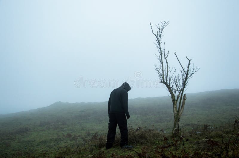 An eerie spooky figure standing by a tree on a foggy winters hillside