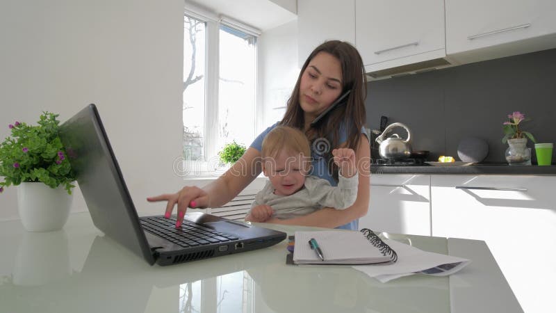 Effort de famille, maman multitâche avec le garçon infantile pleurant tout en travaillant sur l'ordinateur portable et parlant au