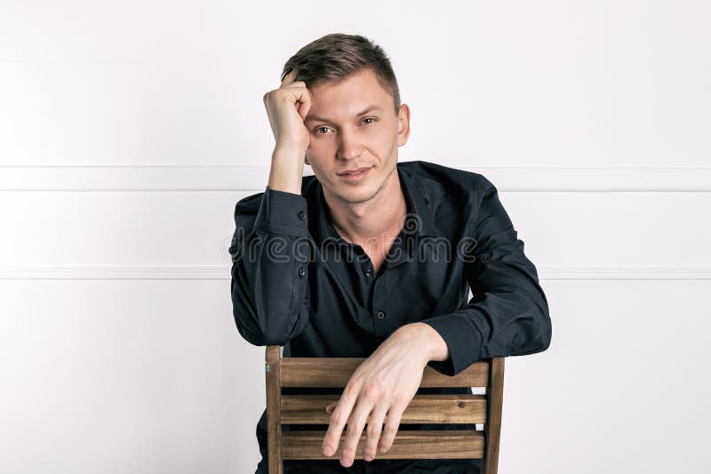 Eftert?nksam ung stilig man i den svarta skjortan som sitter p? stolen och ser kameran royaltyfri bild Eftert?nksam ung stilig man i den svarta skjortan som sitter p? stolen och ser kameran royaltyfri bild