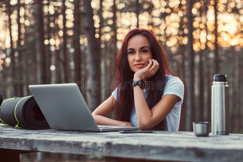 Eftertänksam hipsterflicka i den vita skjortan som ser kameran, medan sitta på en träbänk med en öppen bärbar dator i arkivbilder Eftertänksam hipsterflicka i den vita skjortan som ser kameran, medan sitta på en träbänk med en öppen bärbar dator i arkivbilder