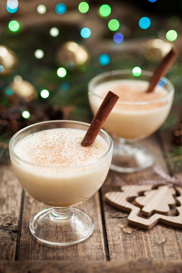 Eggnog traditional christmass egg, milk, rum, vanilla alcoholic liqueur preparation recipe with cinnamon stick on vintage wooden table background. Green and blue bokeh, shallow depth of field. Rustic style.