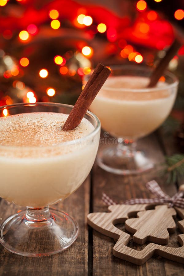 Eggnog traditional homemade christmass celebration egg, milk, rum, vanilla liqueur alcohol drink with cinnamon stick in two glass cups on vintage wooden table. Red bokeh and shallow depth of field. Rustic style.