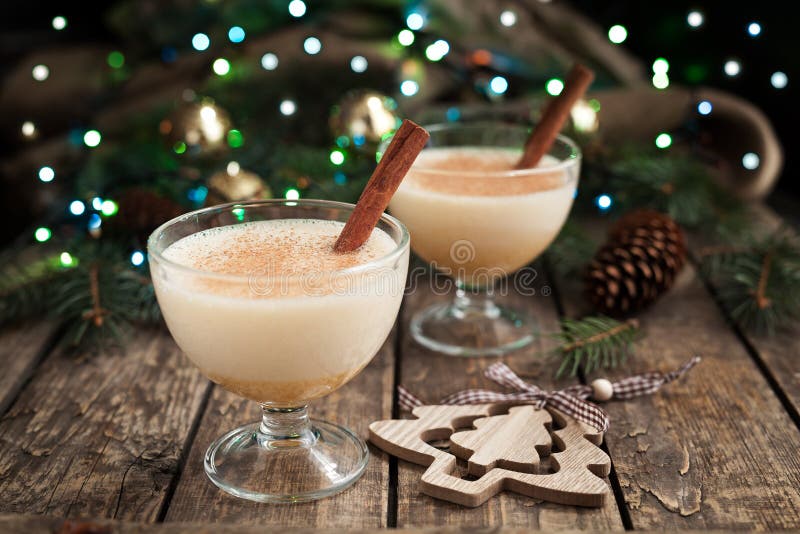 Eggnog traditional xmas homemade winter egg, milk, rum, vanilla alcohol liqueur preparation recipe in two glass cups with cinnamon sticks on wooden vintage table. Blue and green bokeh background. shallow depth of field. Rustic style