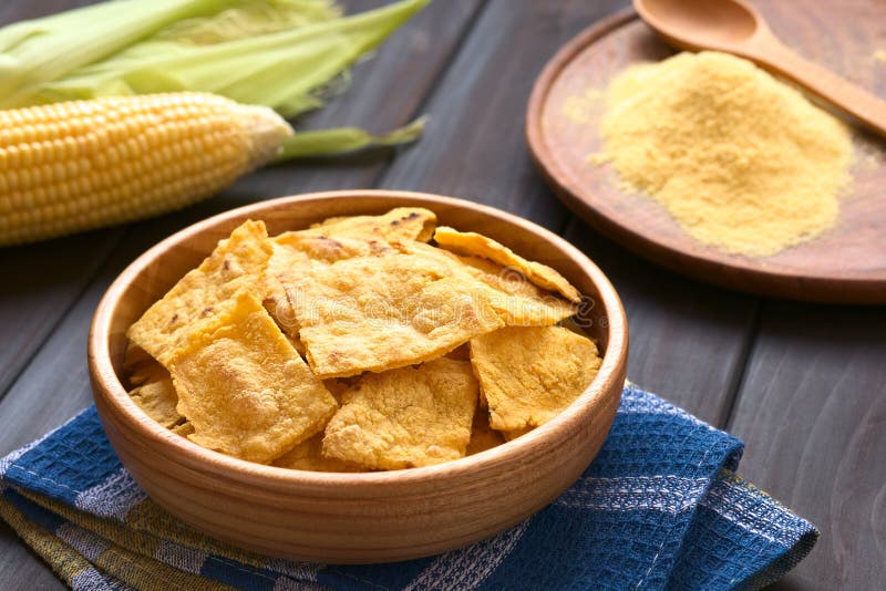 Wooden bowl of homemade baked corn chips with cobs of corn and cornmeal in the back, photographed with natural light (Selective Focus, Focus on the front of the chip on the top). Wooden bowl of homemade baked corn chips with cobs of corn and cornmeal in the back, photographed with natural light (Selective Focus, Focus on the front of the chip on the top)