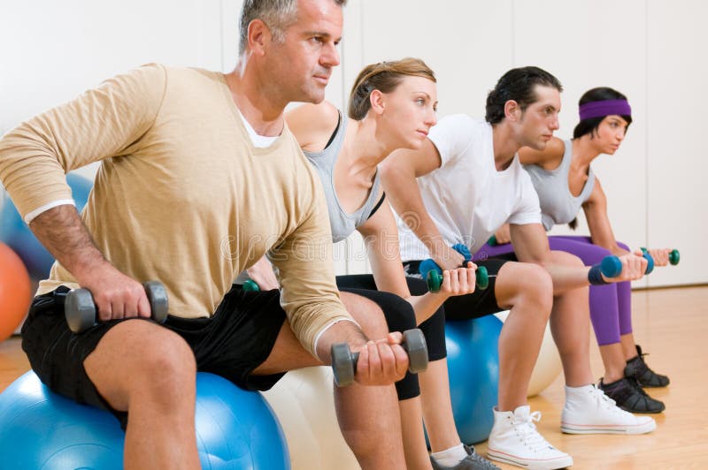 Healthy young people lifting dumbbell and sitting on a fitness ball at gym, instructor on foreground. Healthy young people lifting dumbbell and sitting on a fitness ball at gym, instructor on foreground