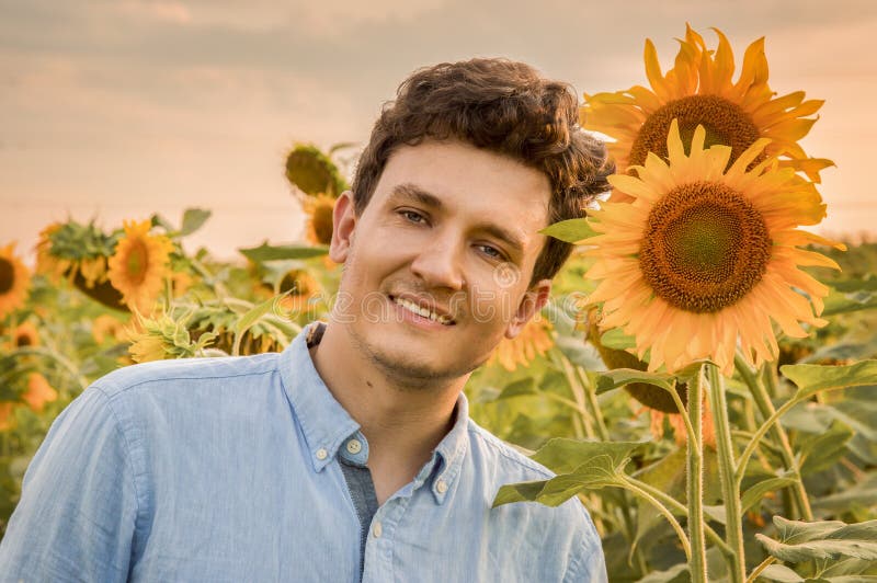 Ein Abschluss herauf Porträt eines jungen lächelnden Mannes auf einem Gebiet von Sonnenblumen Gegen das backdground der untergehe lizenzfreies stockfoto Ein Abschluss herauf Porträt eines jungen lächelnden Mannes auf einem Gebiet von Sonnenblumen Gegen das backdground der untergehe lizenzfreies stockfoto