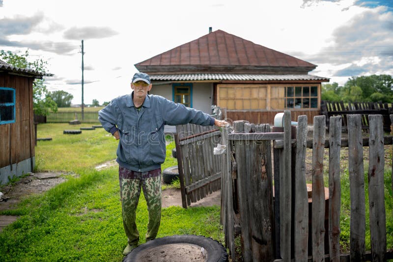 Ein alter Mann in Gläsern und eine Kappe in Hosen mit geschnittenem Gras steht im Dorfhaus lizenzfreies stockbild Ein alter Mann in Gläsern und eine Kappe in Hosen mit geschnittenem Gras steht im Dorfhaus lizenzfreies stockbild