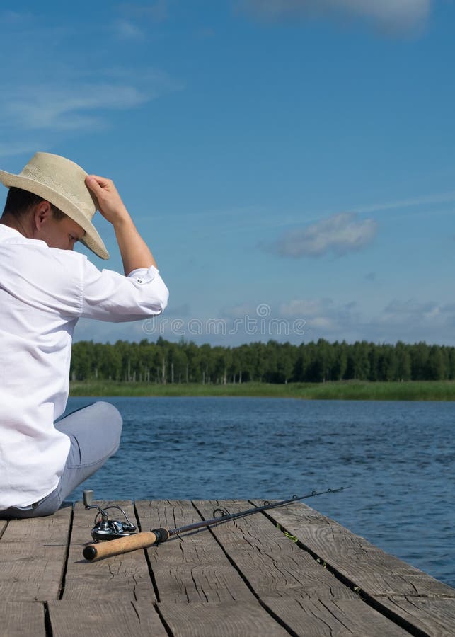 Ein Fischer mit einem Hut sitzt auf dem Pier des Flusses und der Blicke an der Angelrute an einem heißen sonnigen Tag lizenzfreies stockbild Ein Fischer mit einem Hut sitzt auf dem Pier des Flusses und der Blicke an der Angelrute an einem heißen sonnigen Tag lizenzfreies stockbild