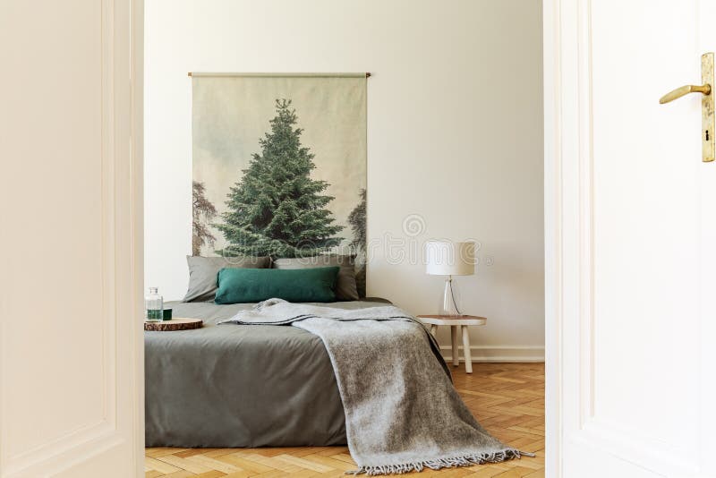 A peek through an open door into a simple style bedroom interior with a bed dressed in gray and green sheets, pillows and a wool blanket. An evergreen tree poster on a background wall. Real photo. concept. A peek through an open door into a simple style bedroom interior with a bed dressed in gray and green sheets, pillows and a wool blanket. An evergreen tree poster on a background wall. Real photo. concept