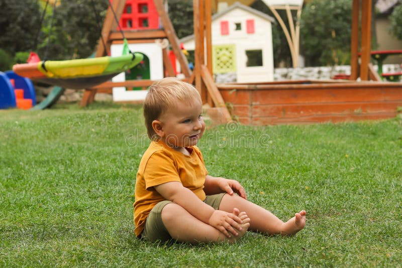 Ein fröhliches Kleinkind, das auf grünem Gras auf einem Spielplatz im Freien sitzt. Das Kind trägt ein orangefarbenes Hemd und Oli stockfotografie