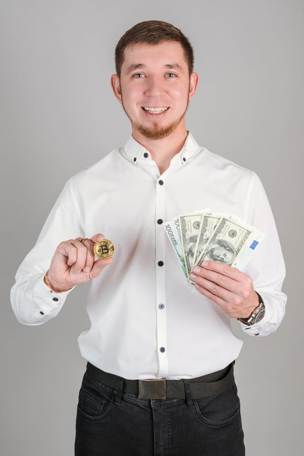 Ein Geschäftsmann in einem weißen Hemd hält eine Bitcoin-Goldmünze und einen Fan von Papier-Dollar-Rechnungen in seiner Hand senkr lizenzfreie stockbilder