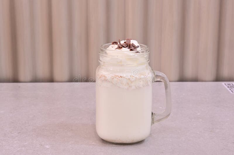 Ein Glas Weißmilch mit Schokoladenpulver oben. Das Glas steht auf dem Tisch.