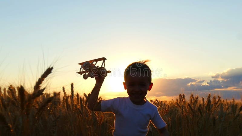 Ein glückliches Kind läuft über ein Weizenfeld während des Sonnenuntergangs und hält eine Spielzeugfläche Der Junge zeigt den Flu