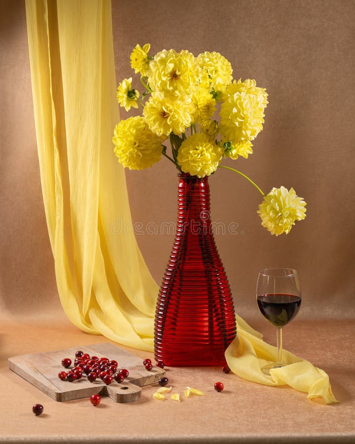 Ein großer Blumenstrauß in roter Vase neben einem Glas Rotwein und roten Beeren oder. Ein großer Blumenstrauß in einer roten Vase neben einem Glas Rotwein und roten Beeren auf orangefarbenem Hintergrund und einem hängenden gelben Schal