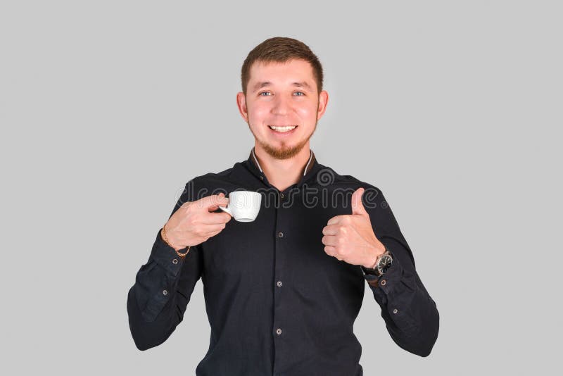 Ein gut aussehender Bärenmann hält eine kleine Kaffeetasse in den Händen und zeigt eine Daumen auf, während er auf einem grauen Hi lizenzfreie stockfotos