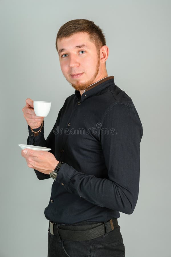 Ein junger, bärtige, rötlich gekleideter Mann trinkt Kaffee aus einem kleinen weißen Krug, der sich die Kamera anschaut.