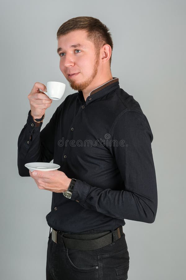 Ein junger bärtige, rötlich-haariger Mann trinkt Kaffee aus einem kleinen weißen Schlamm, der auf einem grau-hintergrundstehenden stockbild
