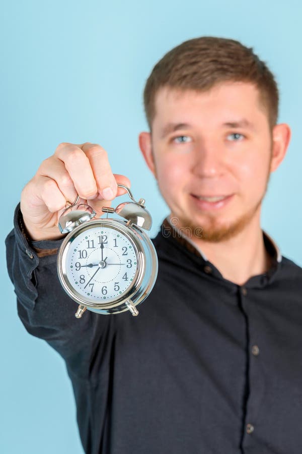 Ein junger erwachsener bärtiger Mann hält einen Wecker mit Waffenlänge, der auf einem selektiven blauen Hintergrund steht stockbild