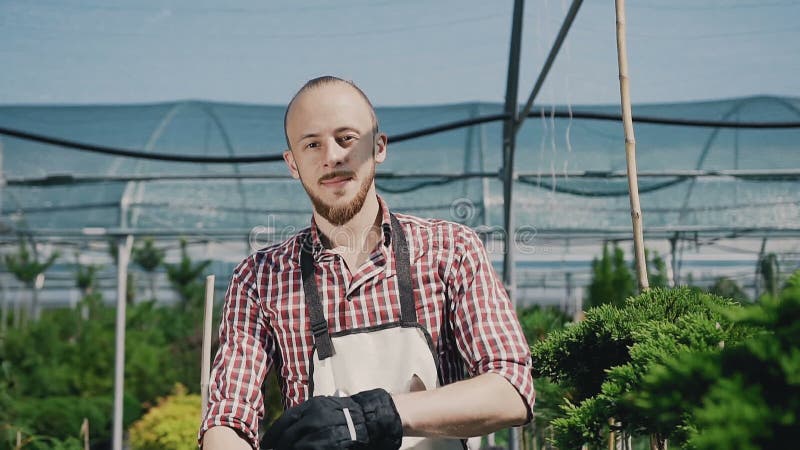 Ein junger Kerl ist ein Gärtner in den Handschuhen und in einem Gartenschutzblech und lächelt und wirft auf der Kamera auf Großes