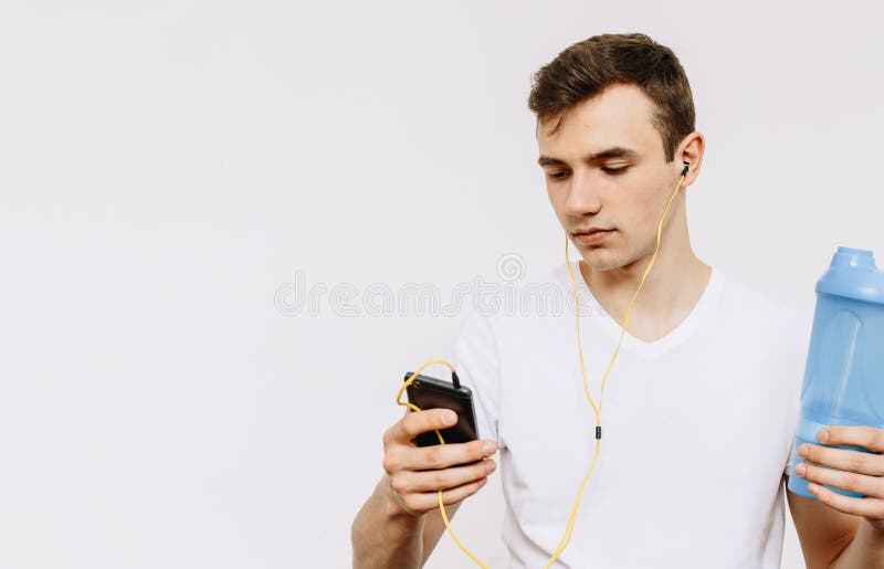 Ein junger Kerl liest Mitteilungen am Telefon und hört Musik auf Kopfhörern und hält einen Schüttel-Apparat in seiner Hand stockfoto Ein junger Kerl liest Mitteilungen am Telefon und hört Musik auf Kopfhörern und hält einen Schüttel-Apparat in seiner Hand stockfoto