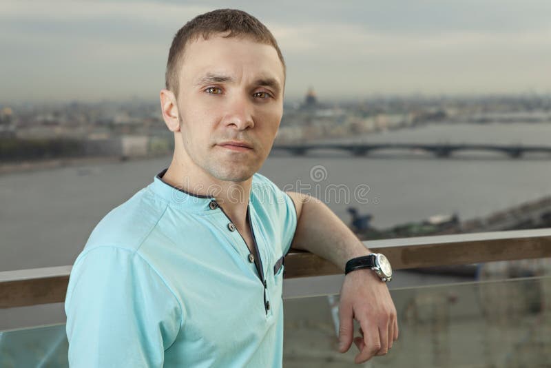 A young man 27 years old, Caucasian, Slav, Russian, dressed in a turquoise shirt with short sleeves, horizontal portrait, close-up, against the background of a panorama of European cities. One person, a male, short hair, outdoor. A young man 27 years old, Caucasian, Slav, Russian, dressed in a turquoise shirt with short sleeves, horizontal portrait, close-up, against the background of a panorama of European cities. One person, a male, short hair, outdoor.