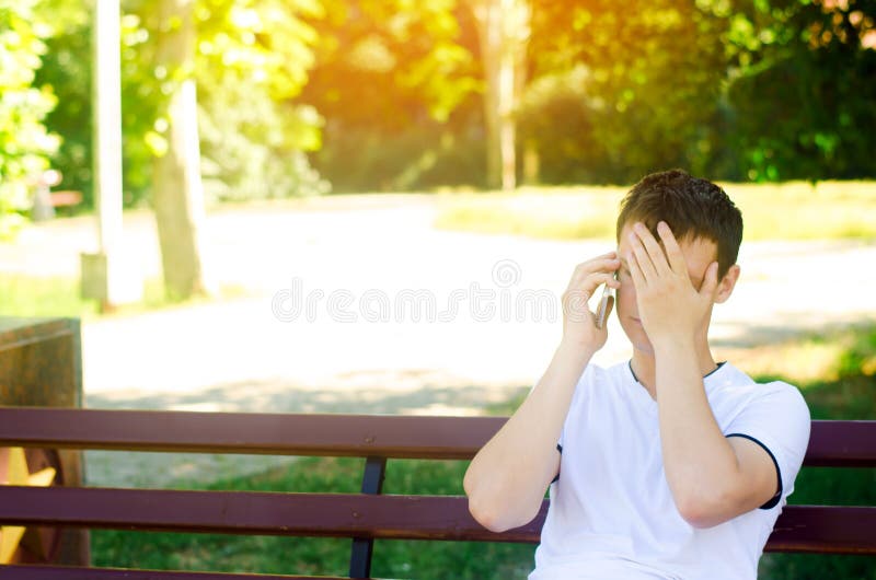 Ein junger nachdenklicher europäischer Kerl in einem weißen T-Shirt spricht am Telefon und sitzt auf einer Bank im Stadtpark bede lizenzfreies stockbild Ein junger nachdenklicher europäischer Kerl in einem weißen T-Shirt spricht am Telefon und sitzt auf einer Bank im Stadtpark bede lizenzfreies stockbild