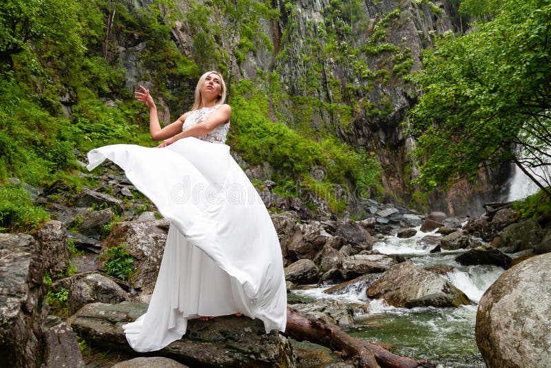 Ein junges blondes Mädchen in einer eleganten Haltung zieht ein Boudoirkleid in den Bergen gegen einen Wasserfall und Steine hoch stockbilder Ein junges blondes Mädchen in einer eleganten Haltung zieht ein Boudoirkleid in den Bergen gegen einen Wasserfall und Steine hoch stockbilder