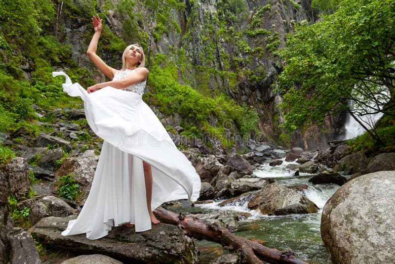 Ein junges blondes Mädchen in einer eleganten Haltung zieht ein Boudoirkleid in den Bergen gegen einen Wasserfall und Steine hoch stockfotos Ein junges blondes Mädchen in einer eleganten Haltung zieht ein Boudoirkleid in den Bergen gegen einen Wasserfall und Steine hoch stockfotos