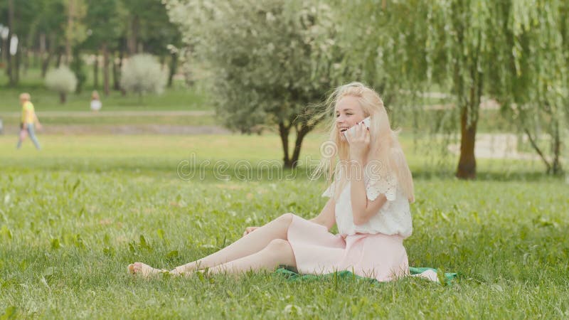 Ein junges blondes Mädchen spricht am Telefon in einem Stadtpark, der auf dem Gras sitzt