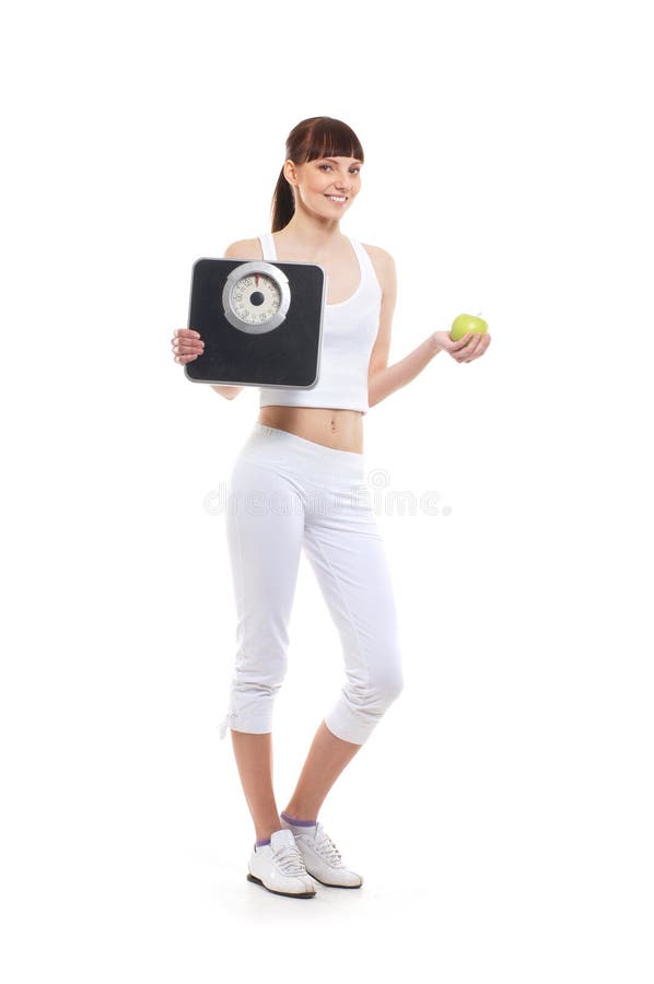 A young and attractive brunette Caucasian woman is holdin a fresh green apple and black weight scales. The image is taken in a studio, isolated on a white background. A young and attractive brunette Caucasian woman is holdin a fresh green apple and black weight scales. The image is taken in a studio, isolated on a white background.
