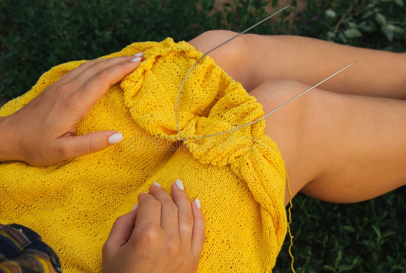 a young girl with long blond hair knits a yellow sweater in the garden in the summer. woman makes clothes with hands closeup. a young girl with long blond hair knits a yellow sweater in the garden in the summer. woman makes clothes with hands closeup.