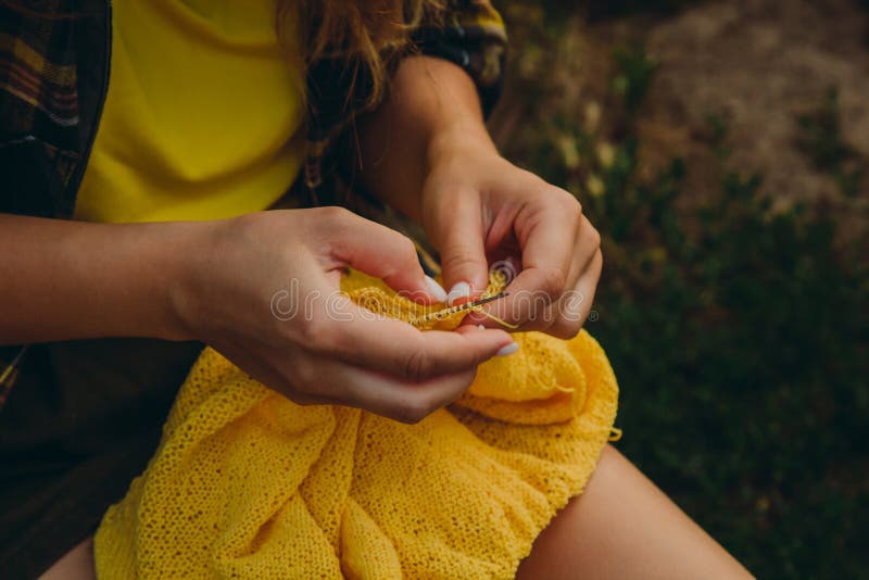 A young girl with long blond hair knits a yellow sweater in the garden in the summer. woman makes clothes with hands closeup. A young girl with long blond hair knits a yellow sweater in the garden in the summer. woman makes clothes with hands closeup.